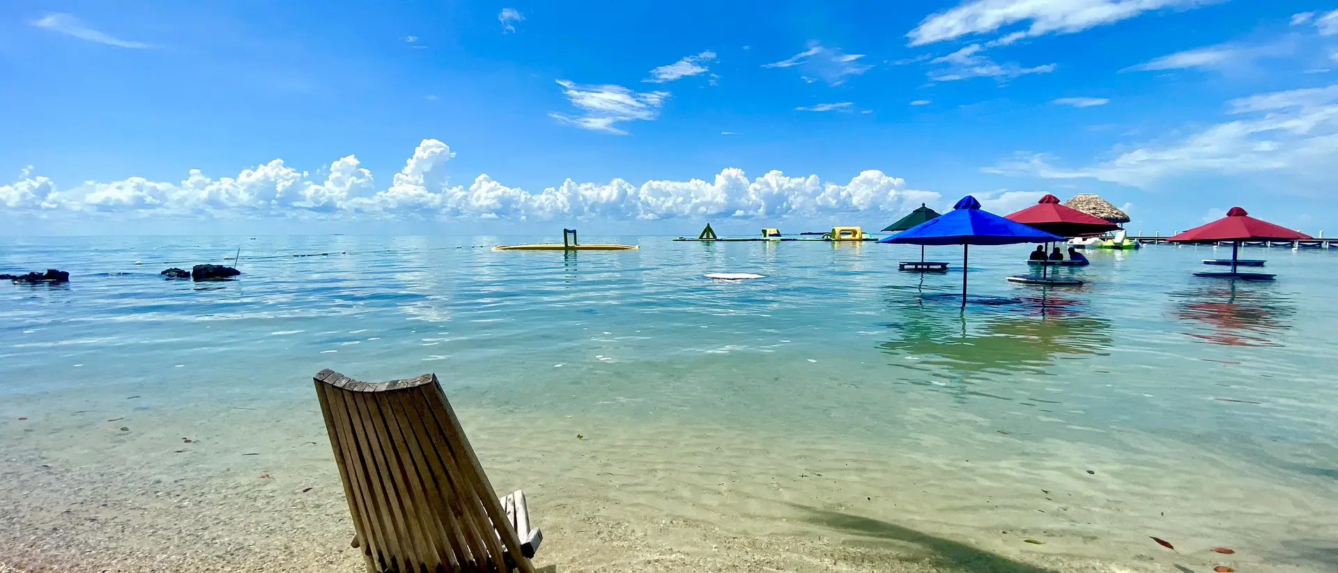 Beach scene with a wooden lounge chair, clear water, and colorful umbrellas under a bright blue sky.