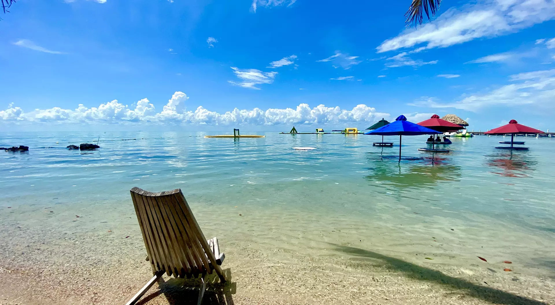 Beach scene with a wooden lounge chair, clear water, and colorful umbrellas under a bright blue sky.