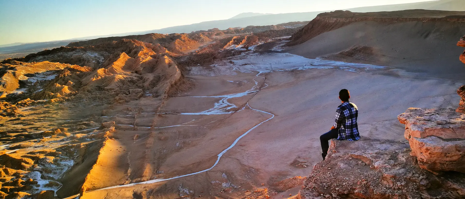 A person sits on a rocky ledge, overlooking a vast desert landscape at sunset.