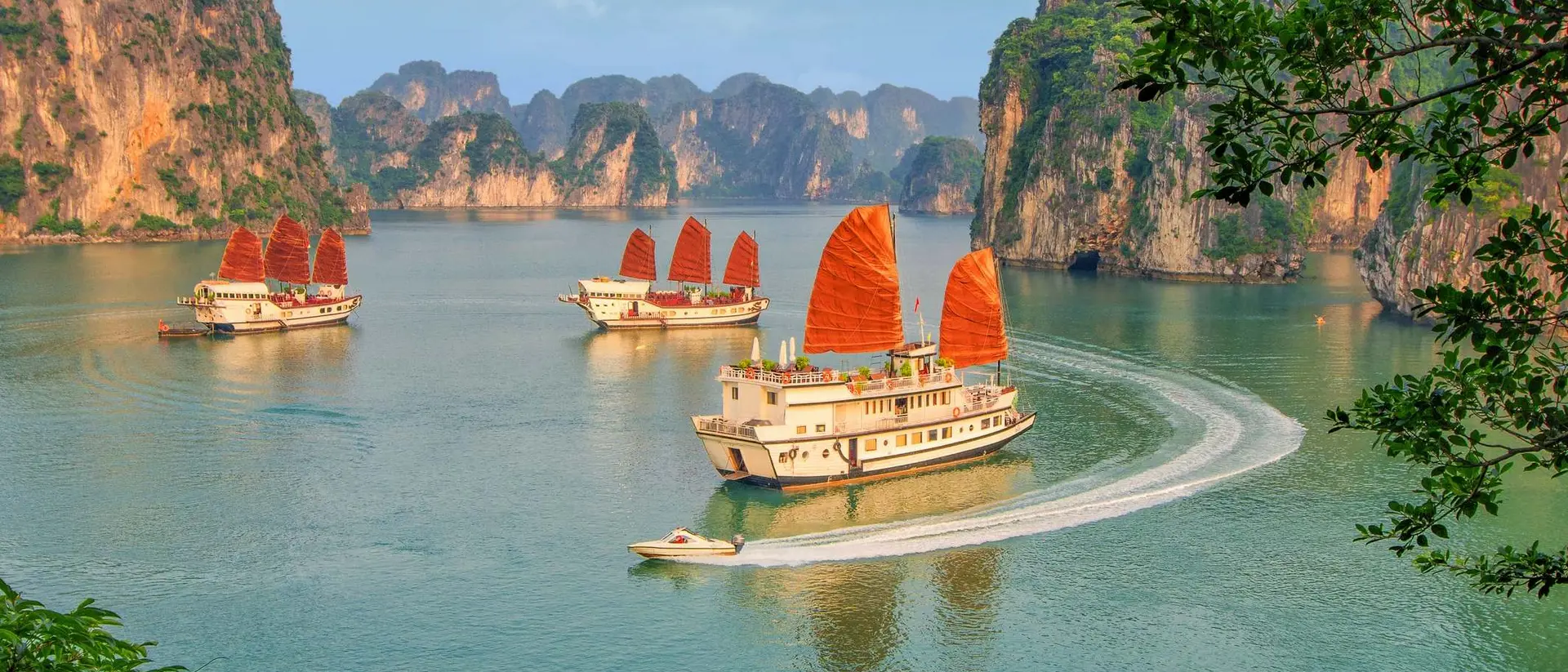 Three traditional wooden boats with red sails glide through a serene bay surrounded by lush green cliffs.