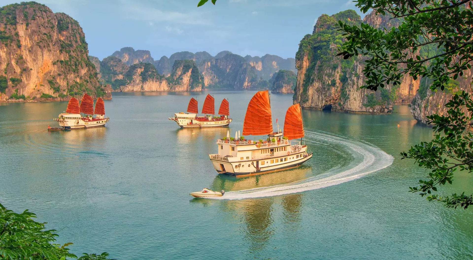 Three traditional wooden boats with red sails glide through a serene bay surrounded by lush green cliffs.
