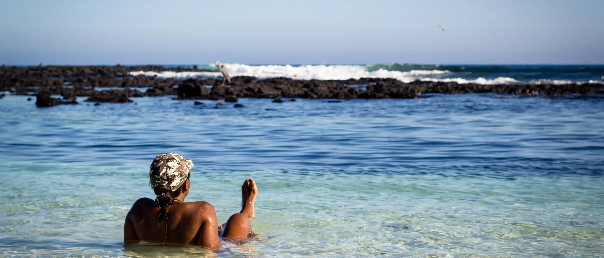 A person relaxes in shallow water at a beach, facing the ocean and surrounded by rocky shoreline under a clear sky.