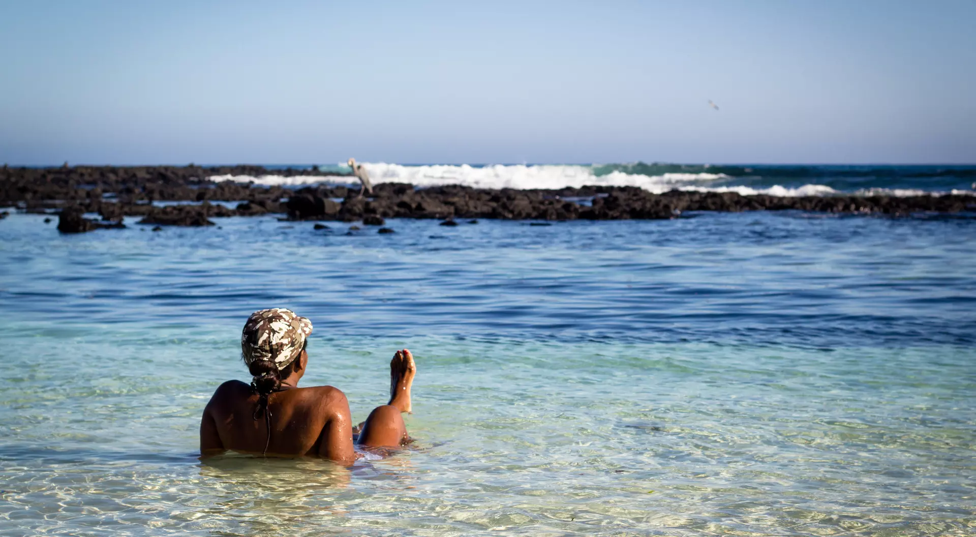 A person relaxes in shallow water at a beach, facing the ocean and surrounded by rocky shoreline under a clear sky.
