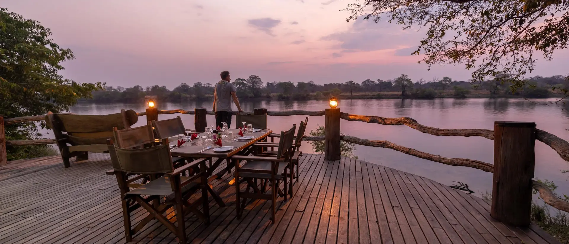 A wooden deck by a river at sunset, with a dining table set and a person standing in the background.