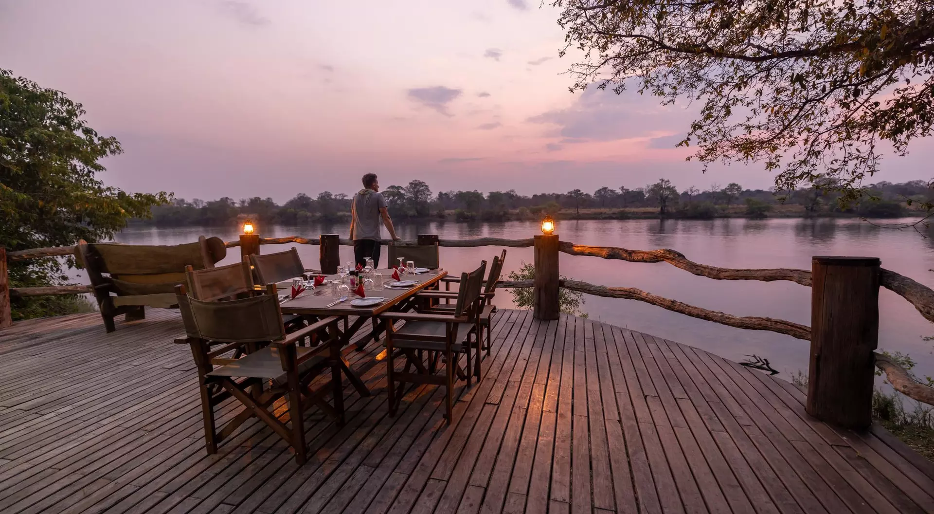A wooden deck by a river at sunset, with a dining table set and a person standing in the background.