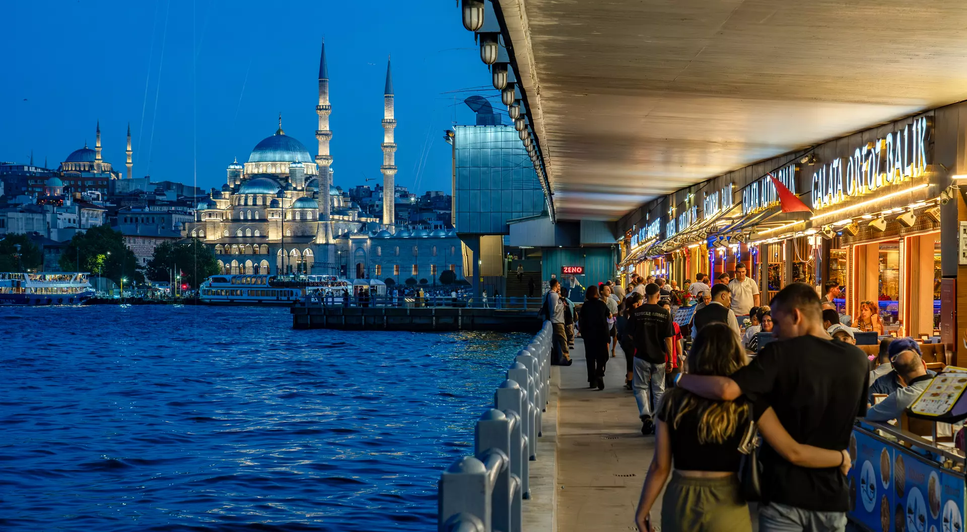 A bustling riverside promenade at dusk, with people walking and cafes, and a view of a mosque in the background.