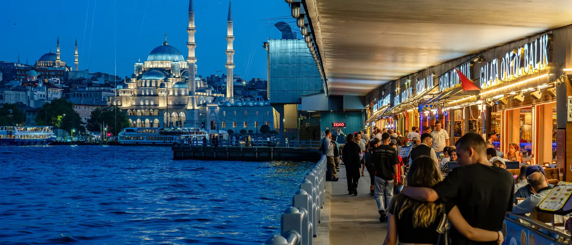 A bustling riverside promenade at dusk, with people walking and cafes, and a view of a mosque in the background.