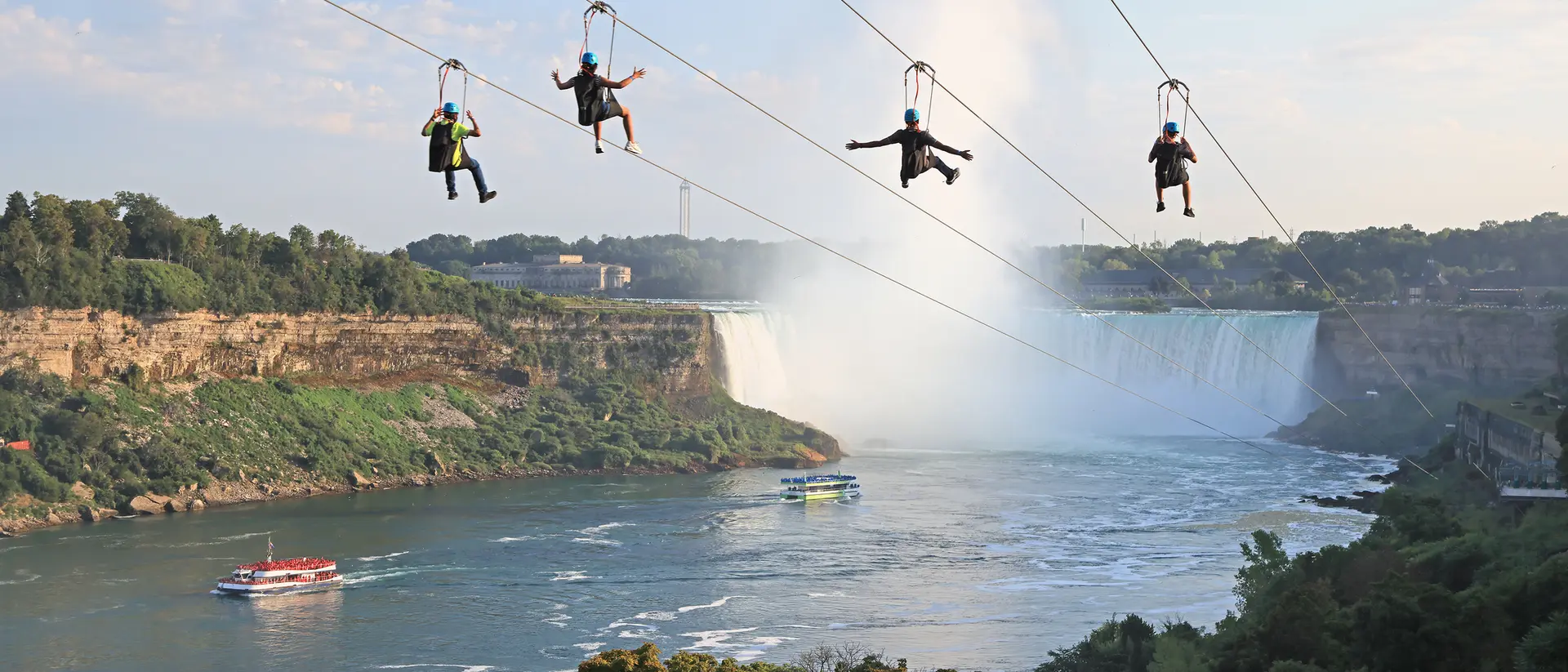 Four people zip-lining over a river near a waterfall, with lush greenery and a boat visible below.
