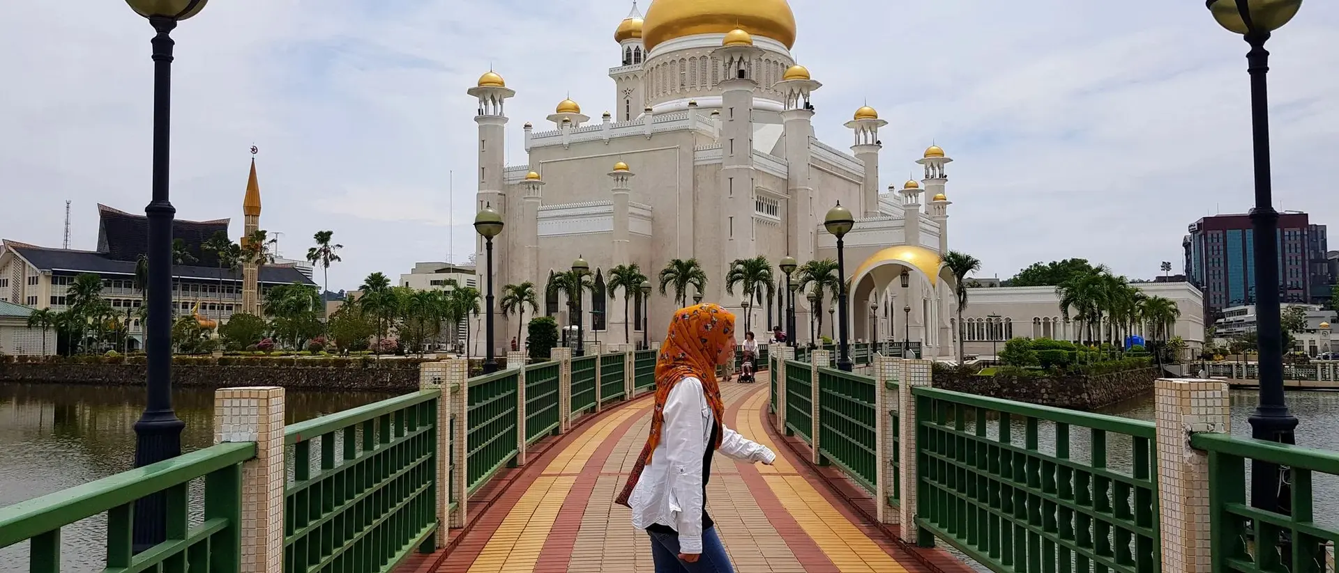 A person with orange hair walks on a bridge toward a grand mosque with golden domes, surrounded by water.