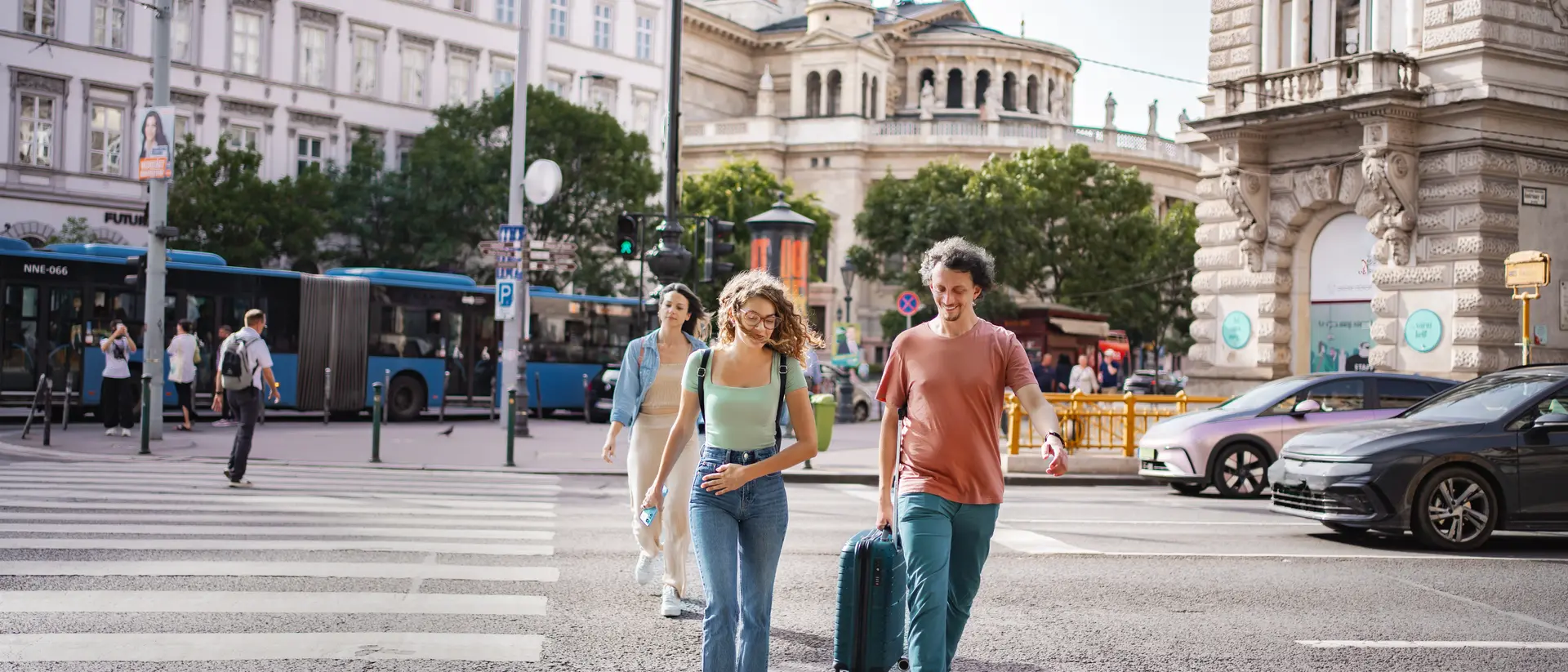 A group of three people walk across a street with a suitcase, surrounded by city buildings and buses.