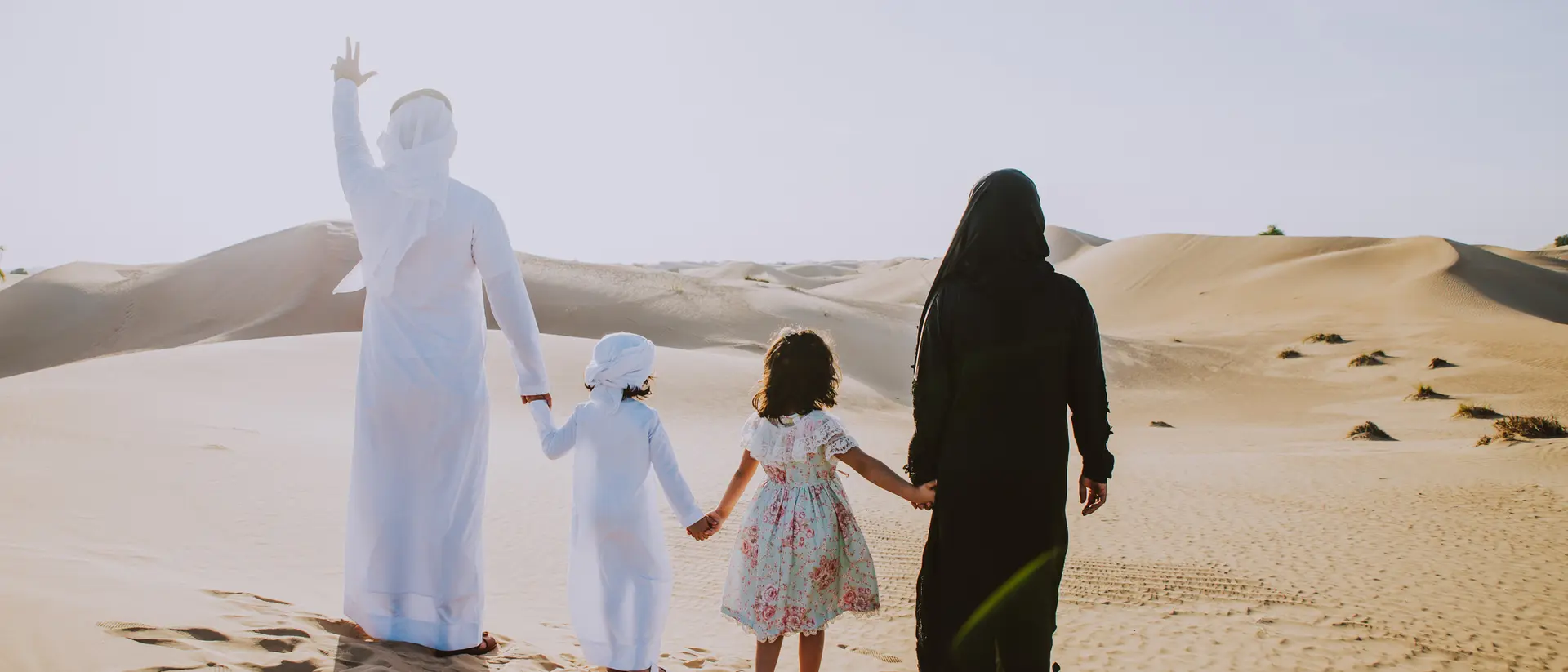 A family of four walks hand in hand through a sandy desert, with one person waving and sunlight shining bright.