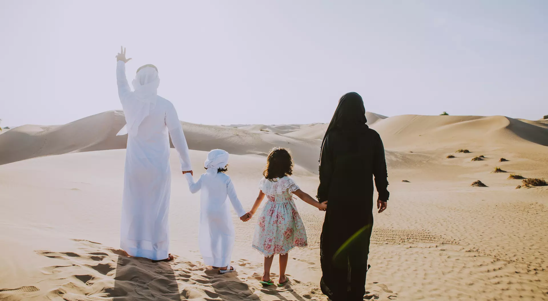 A family of four walks hand in hand through a sandy desert, with one person waving and sunlight shining bright.
