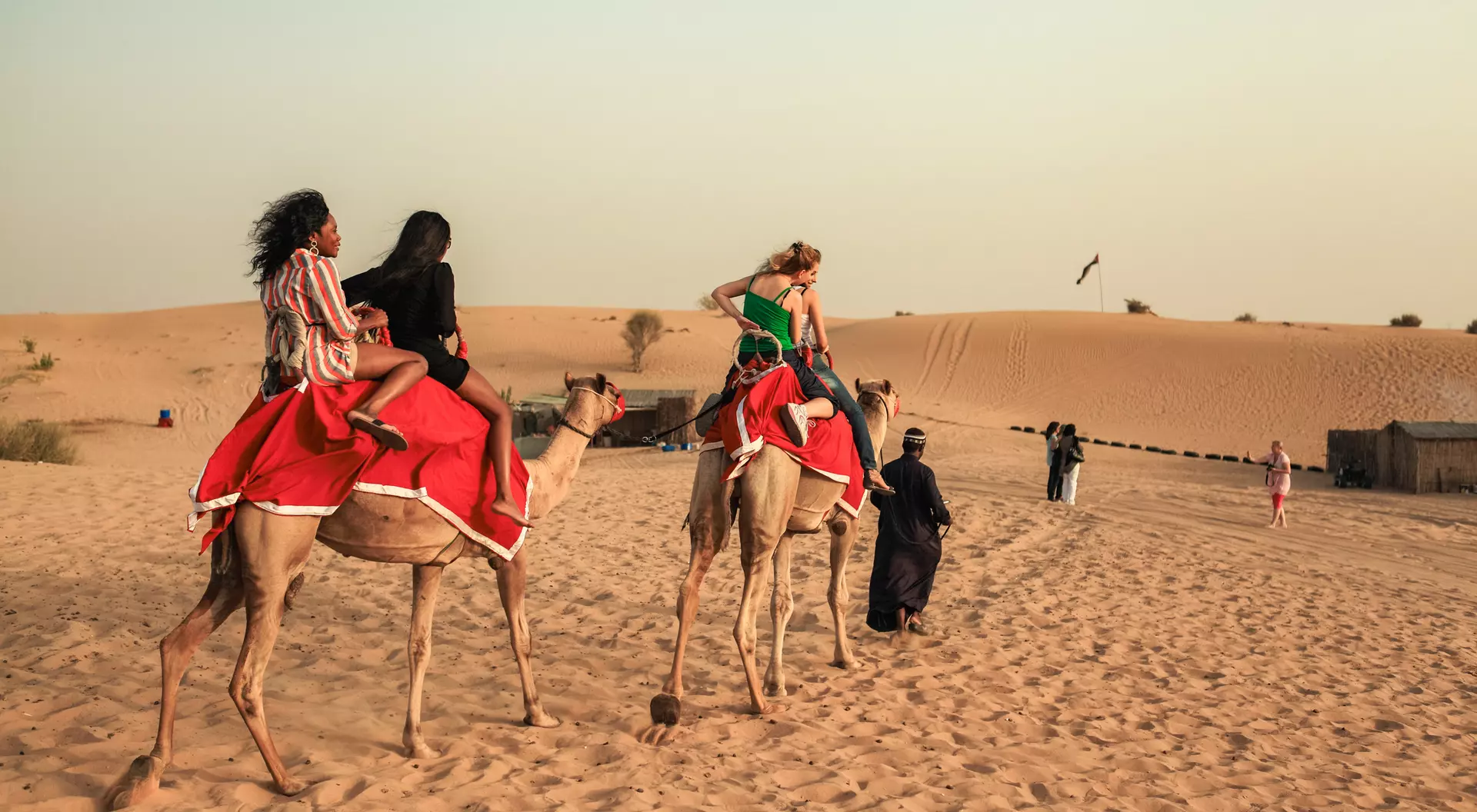Two people riding camels in the desert, with sand dunes in the background and others walking nearby.