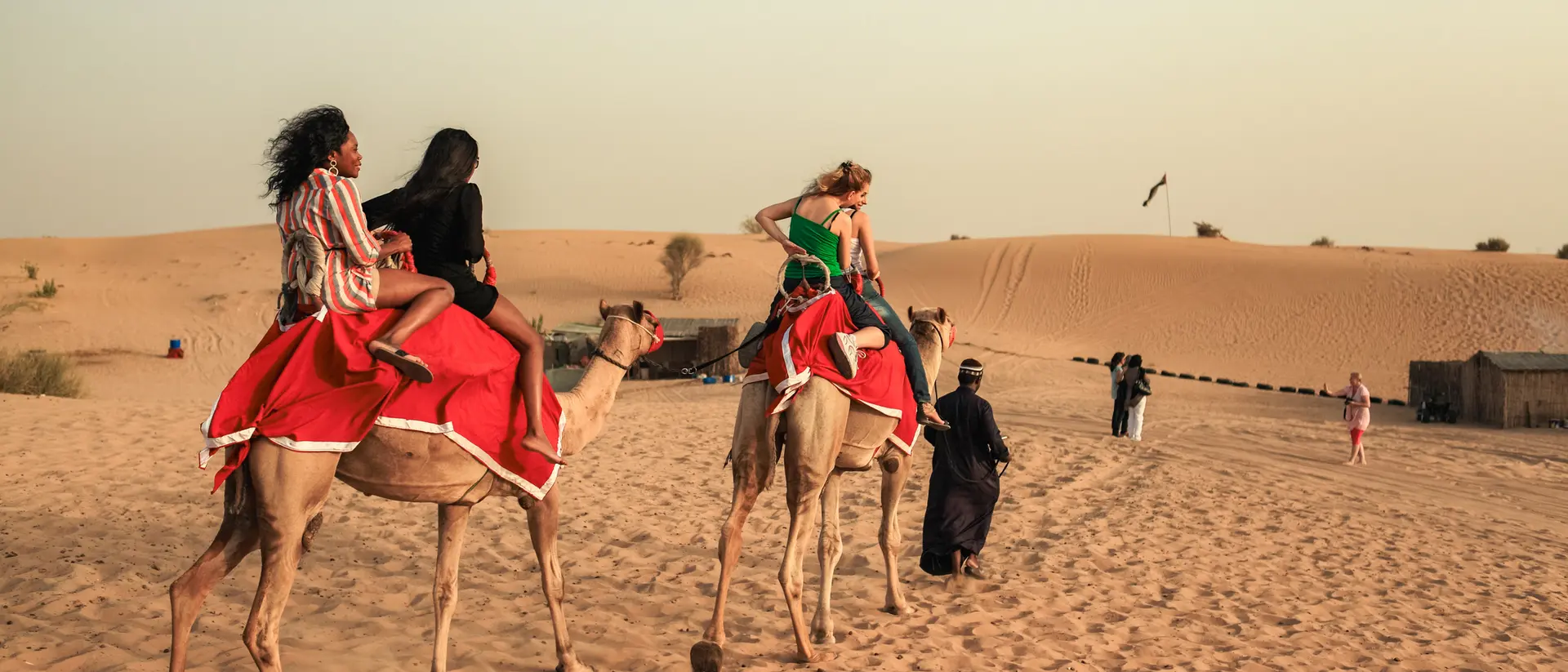 Two people riding camels in the desert, with sand dunes in the background and others walking nearby.