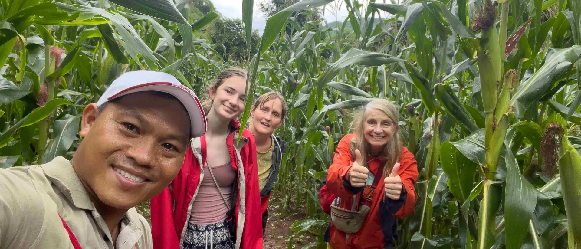 A group of four people smiling in a cornfield, surrounded by tall corn plants on a cloudy day.