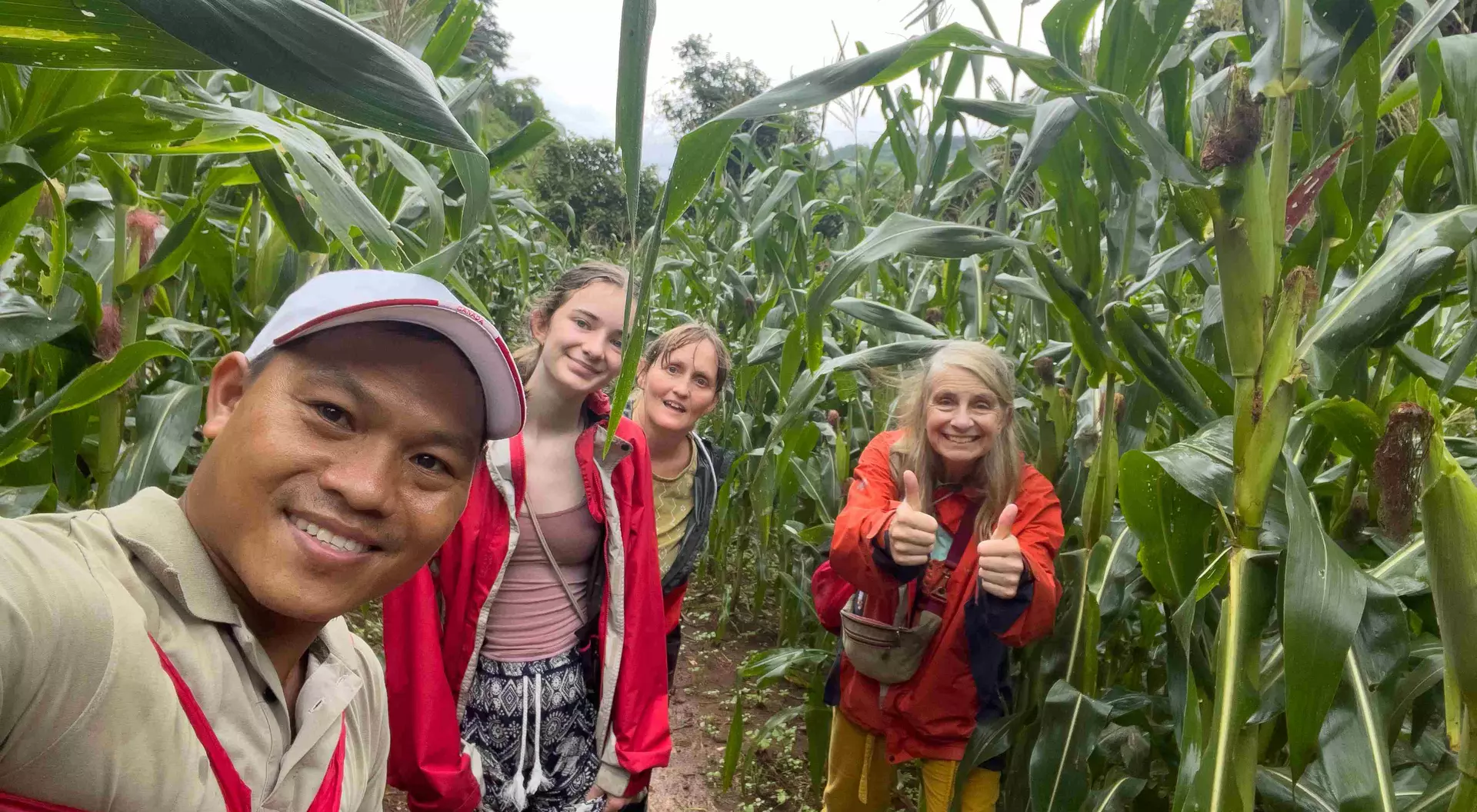 A group of four people smiling in a cornfield, surrounded by tall corn plants on a cloudy day.