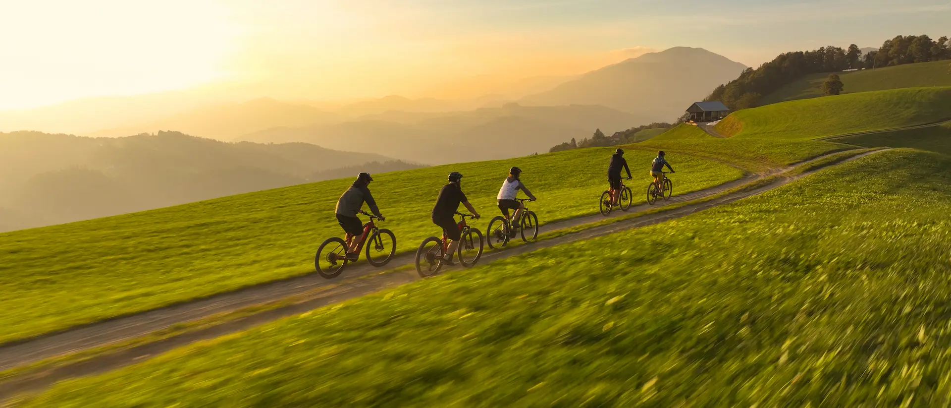 A group of six cyclists riding on a dirt path through a green field at sunset, with mountains in the background.
