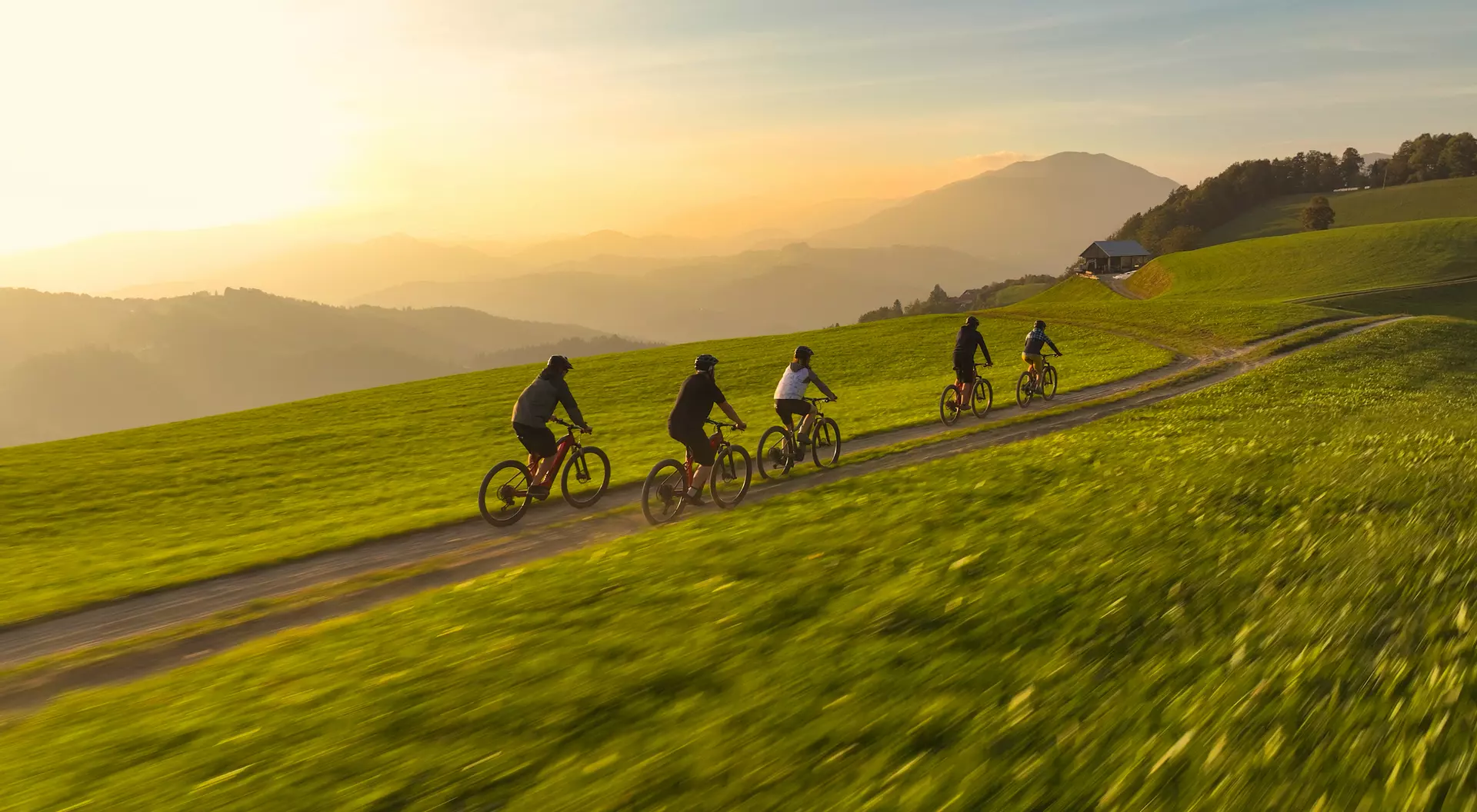 A group of six cyclists riding on a dirt path through a green field at sunset, with mountains in the background.