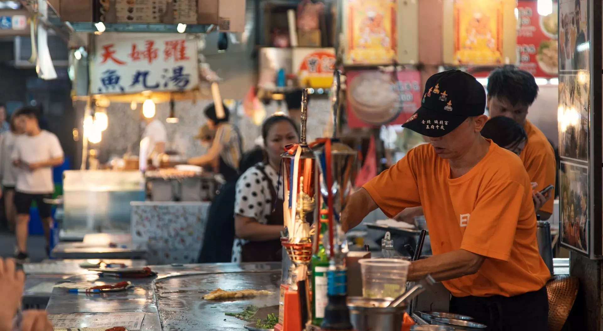 A busy food stall with people, a vendor in an orange shirt cooking, and a woman in polka dots at a table.