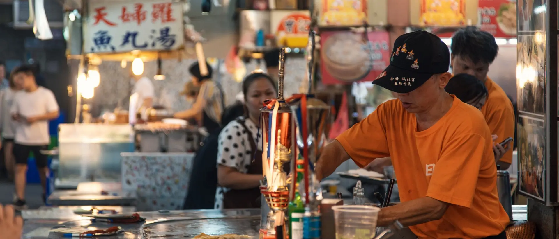 A busy food stall with people, a vendor in an orange shirt cooking, and a woman in polka dots at a table.