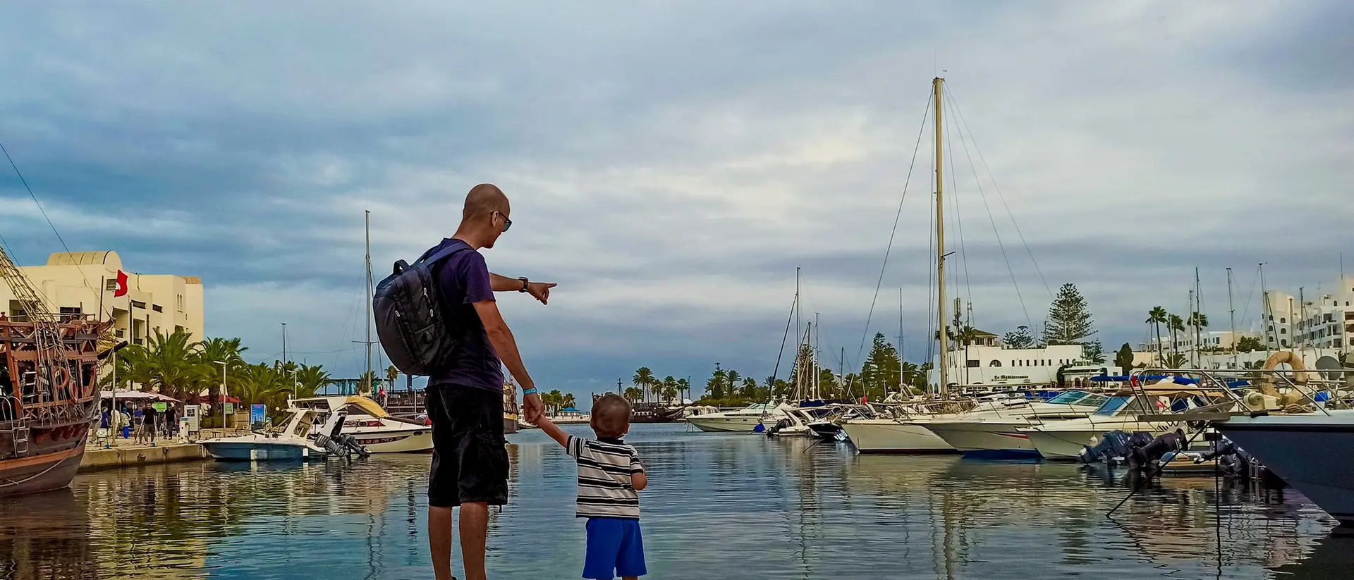 A man and a child stand by a marina, pointing at boats against a cloudy sky.