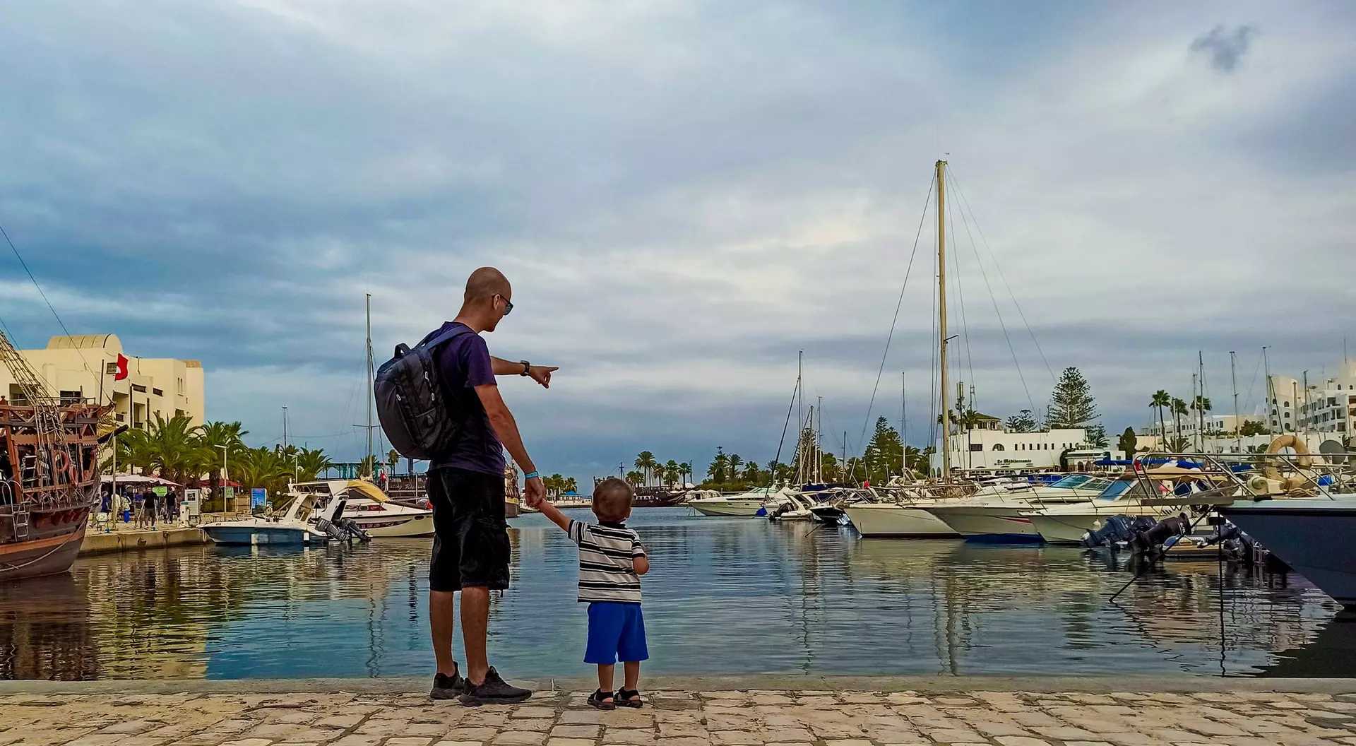 A man and a child stand by a marina, pointing at boats against a cloudy sky.