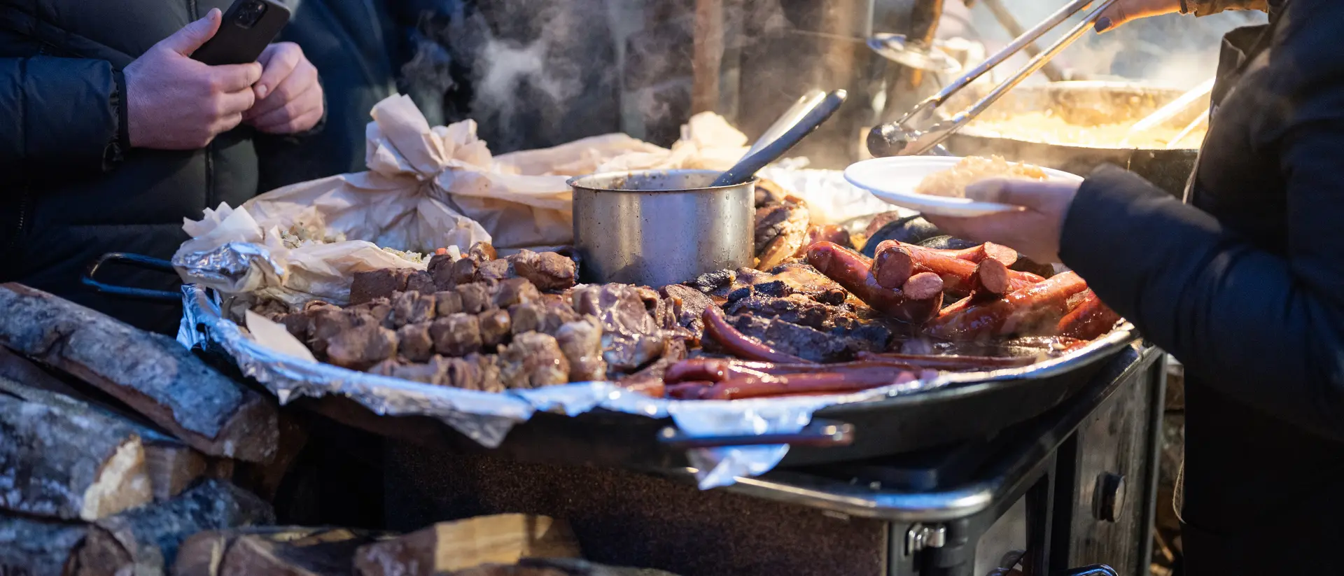 Close-up of a food stall with various meats being served, steam rising, and people in the background.