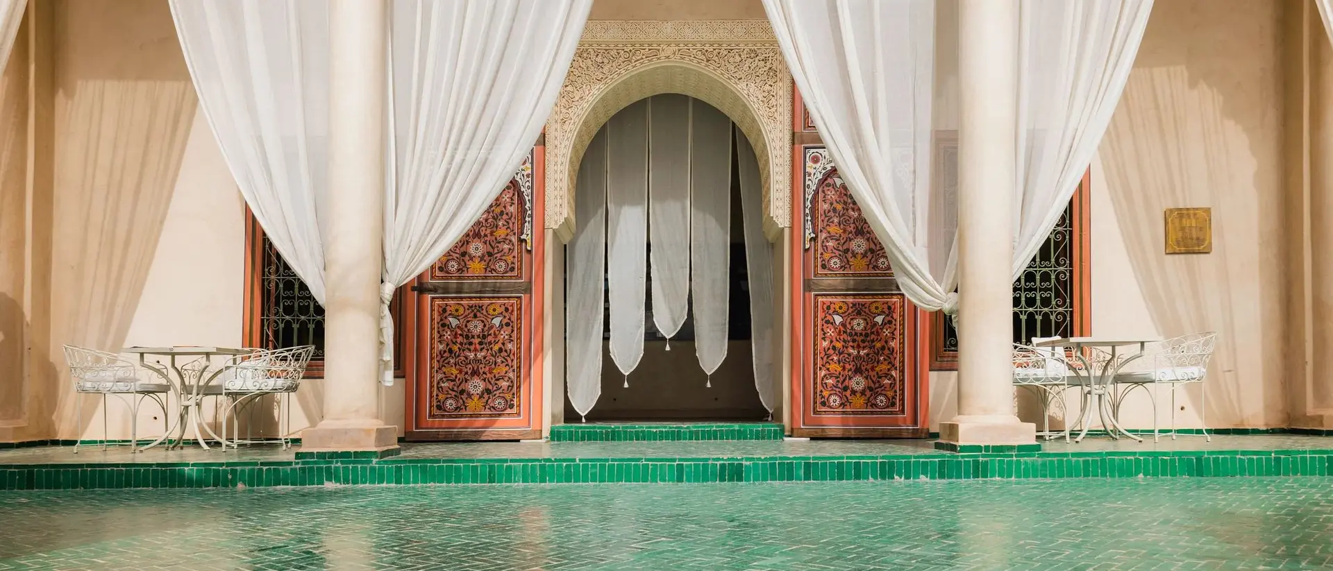 Elegant interior with green tiled floor, sheer drapes, and intricately carved wooden doors in a Moroccan-style setting.
