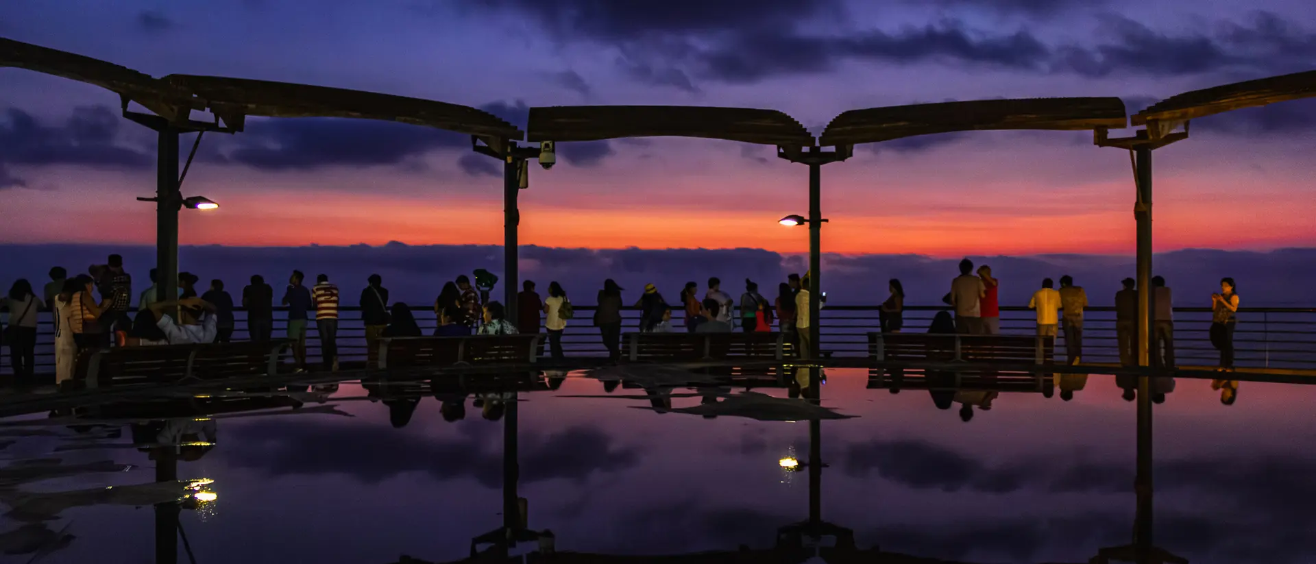Silhouetted people on a pier at sunset, with vibrant orange and purple sky reflected in water below.