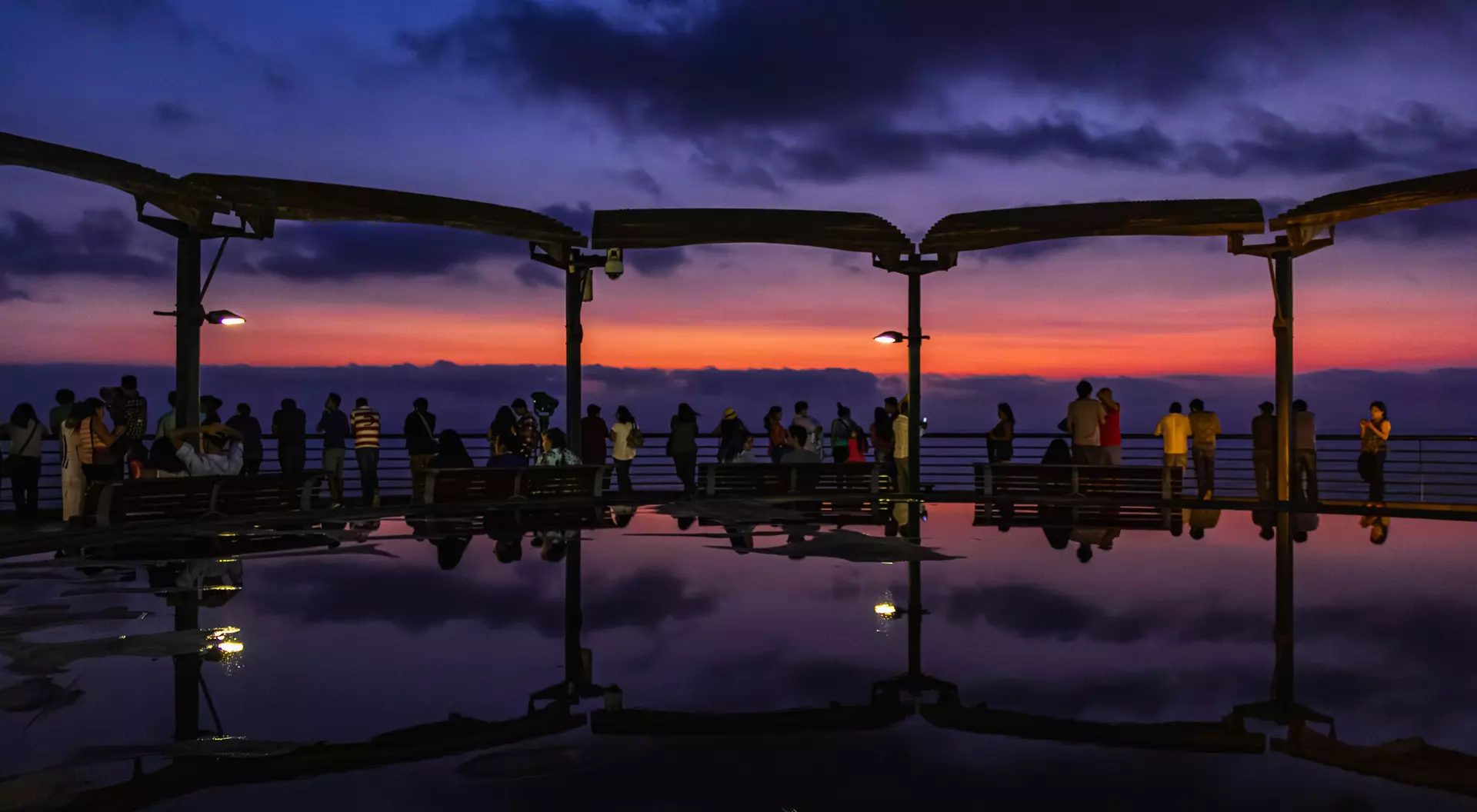 Silhouetted people on a pier at sunset, with vibrant orange and purple sky reflected in water below.