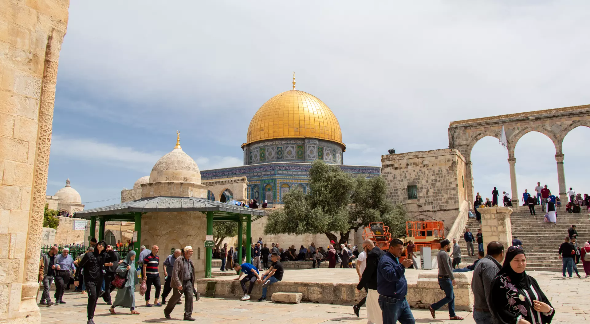 Crowds gather at the Al-Aqsa Mosque compound, featuring the iconic golden dome under a partly cloudy sky.