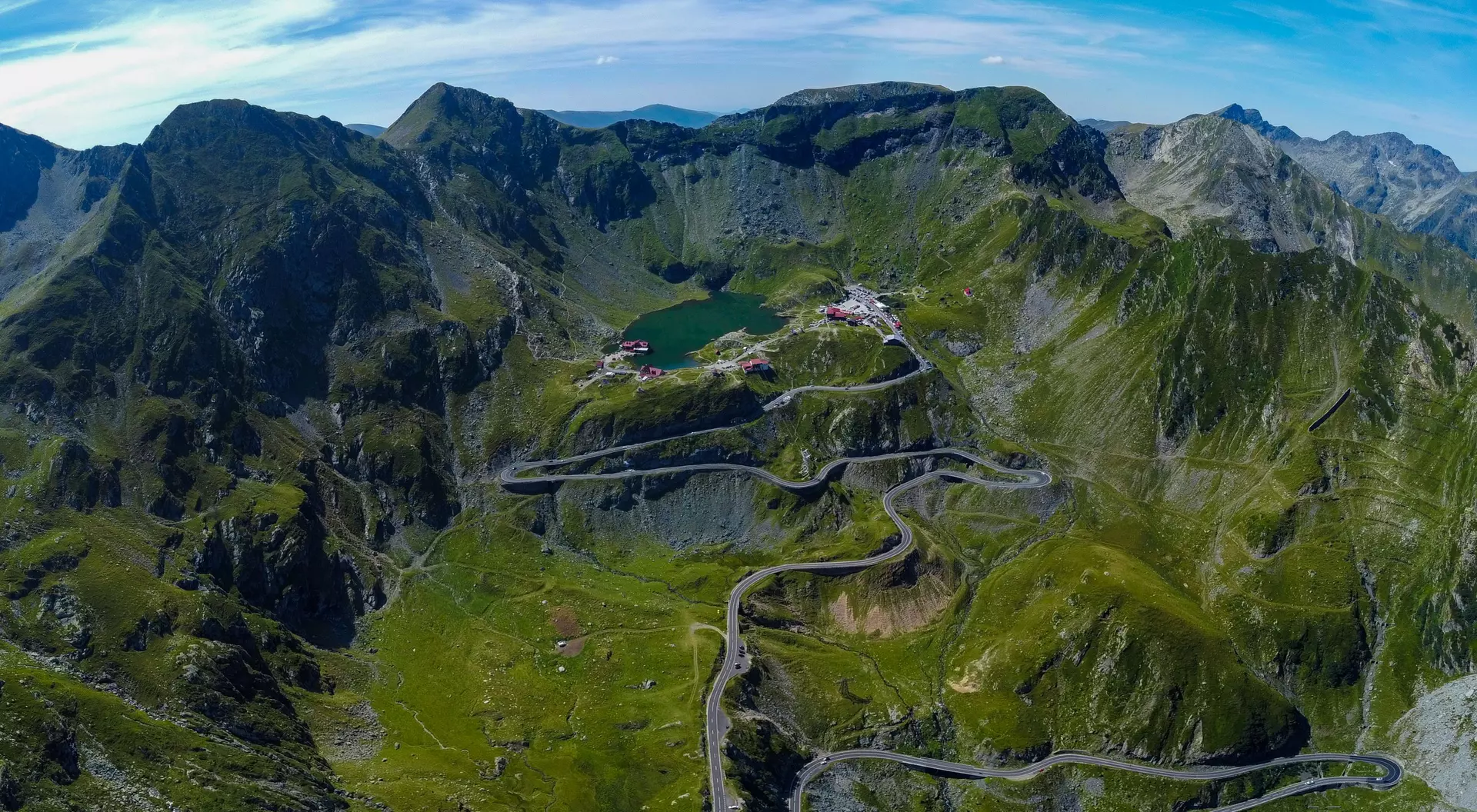 Aerial view of a mountainous landscape with winding roads, a green lake, and lush greenery under a bright blue sky.