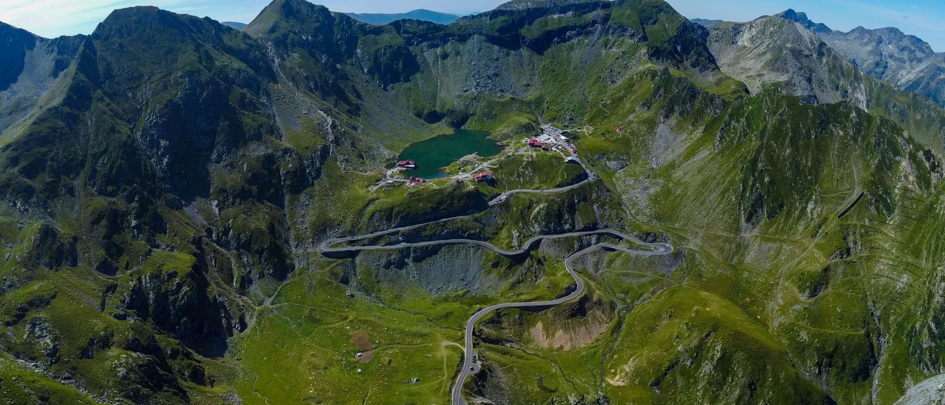 Aerial view of a mountainous landscape with winding roads, a green lake, and lush greenery under a bright blue sky.