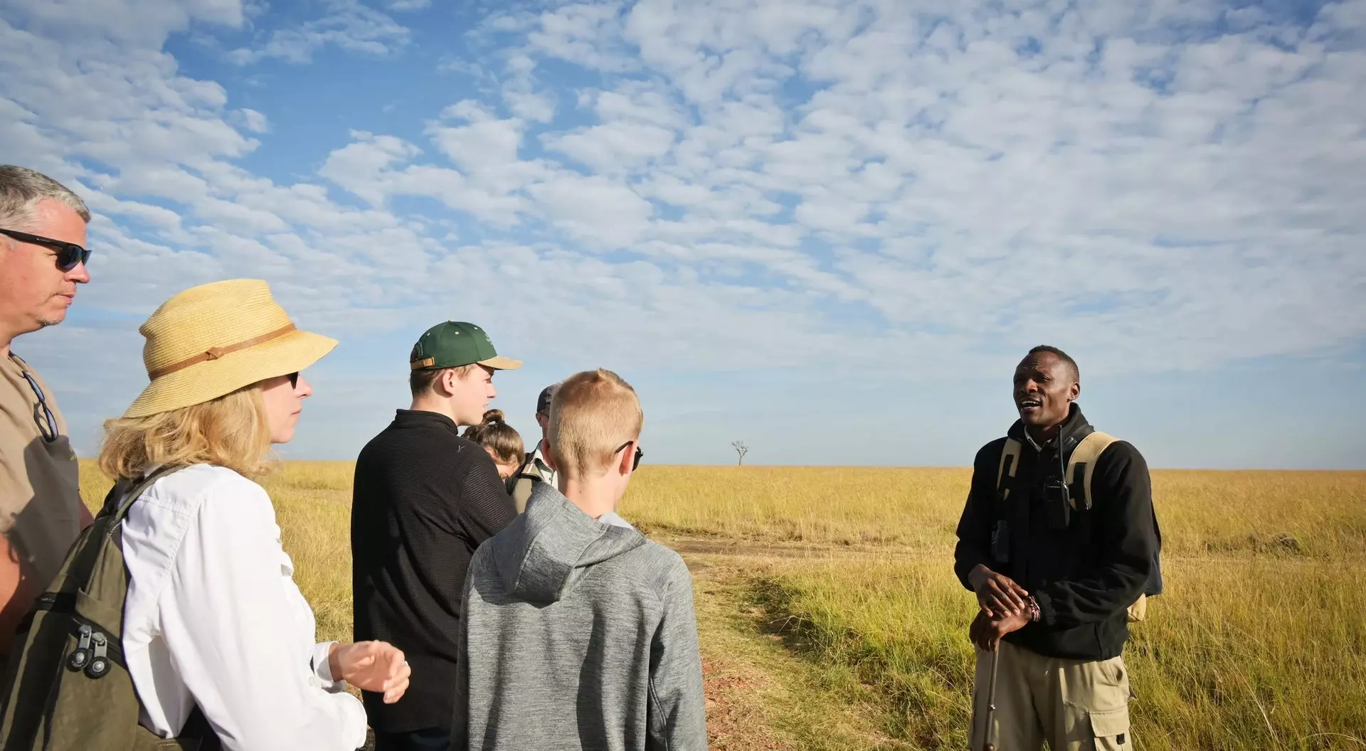 A diverse group of six people listens to a guide in a grassy field under a blue sky with scattered clouds.