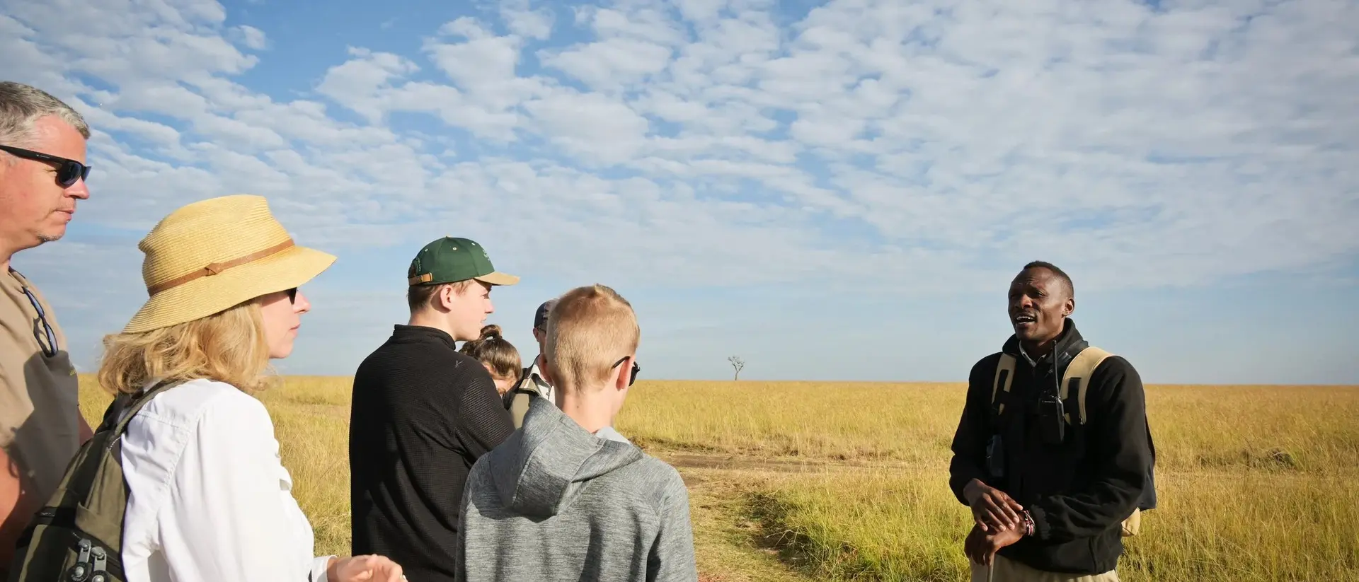 A diverse group of six people listens to a guide in a grassy field under a blue sky with scattered clouds.