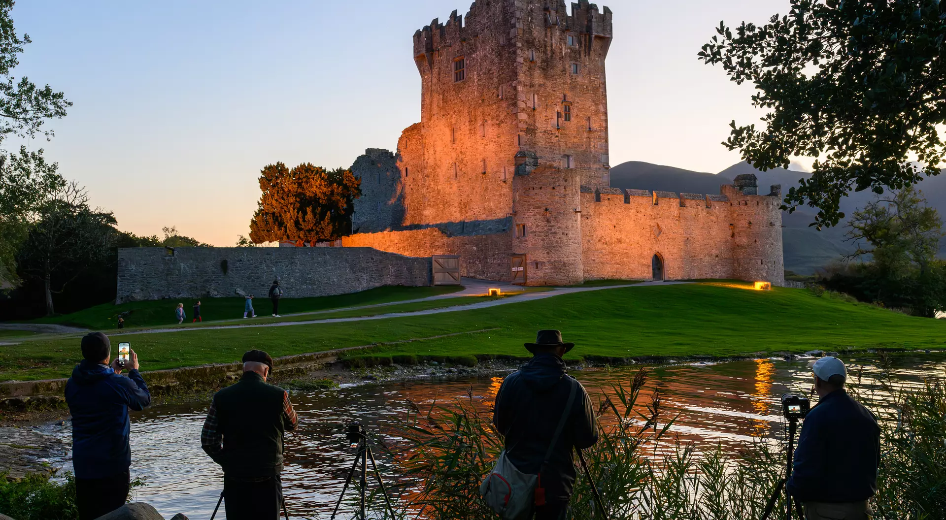 Four photographers capture a castle at sunset, reflected in a serene river, surrounded by lush greenery.