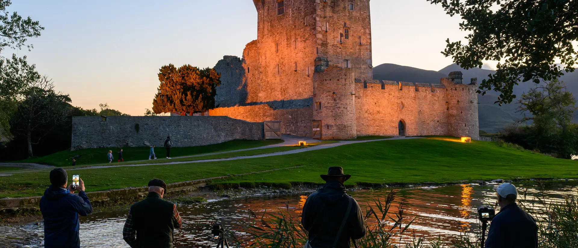Four photographers capture a castle at sunset, reflected in a serene river, surrounded by lush greenery.