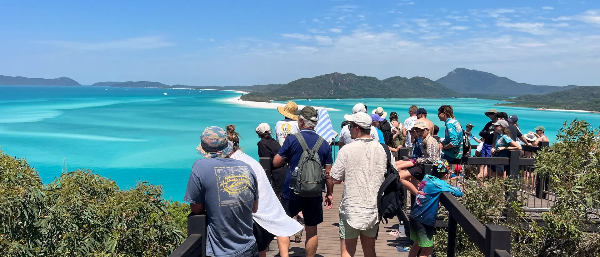 A crowd of people stands on a viewing platform overlooking a vibrant turquoise ocean and distant islands.