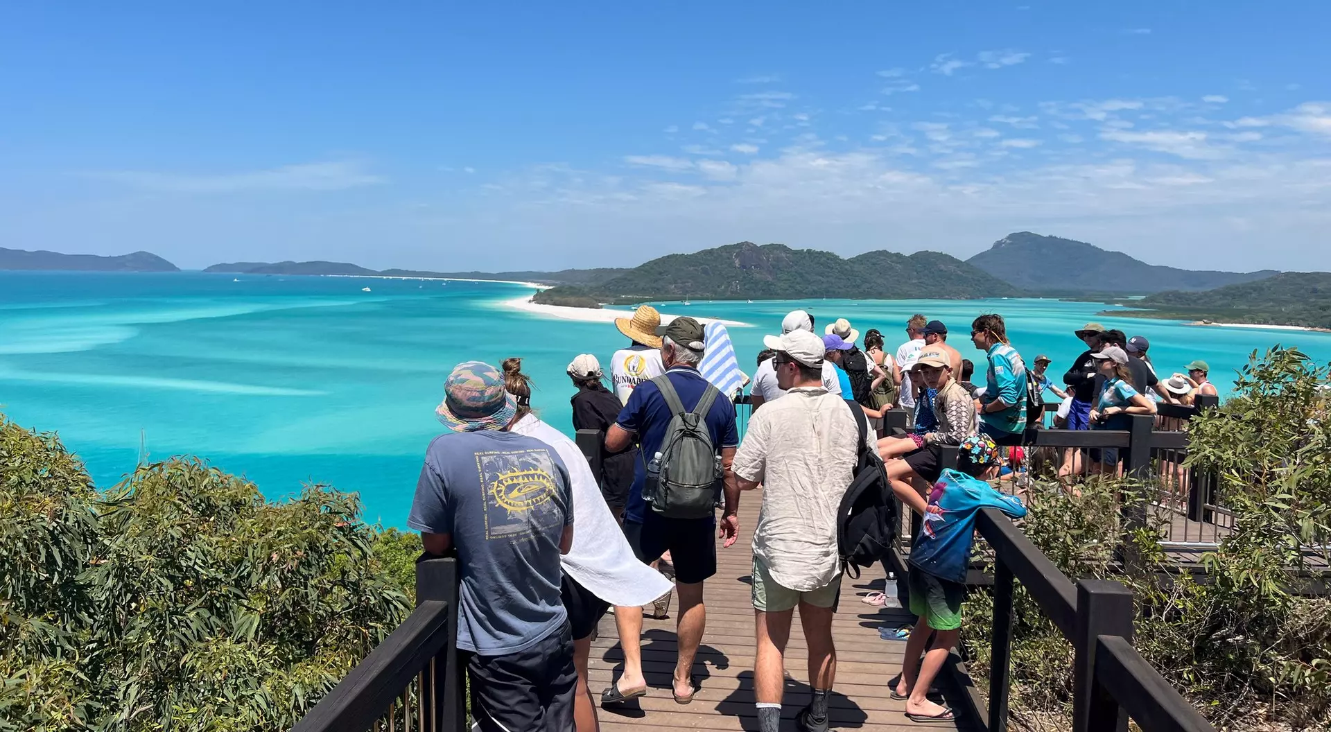 A crowd of people stands on a viewing platform overlooking a vibrant turquoise ocean and distant islands.