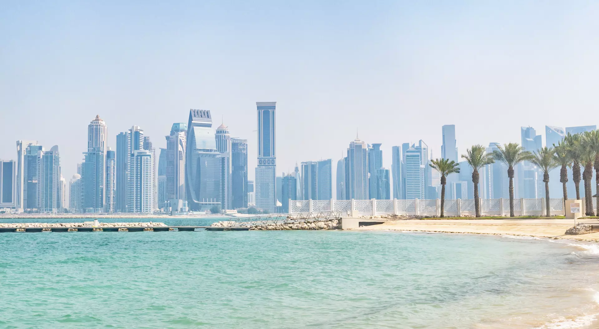 View of a sandy beach with palm trees and a skyline of tall buildings in the background under a clear sky.