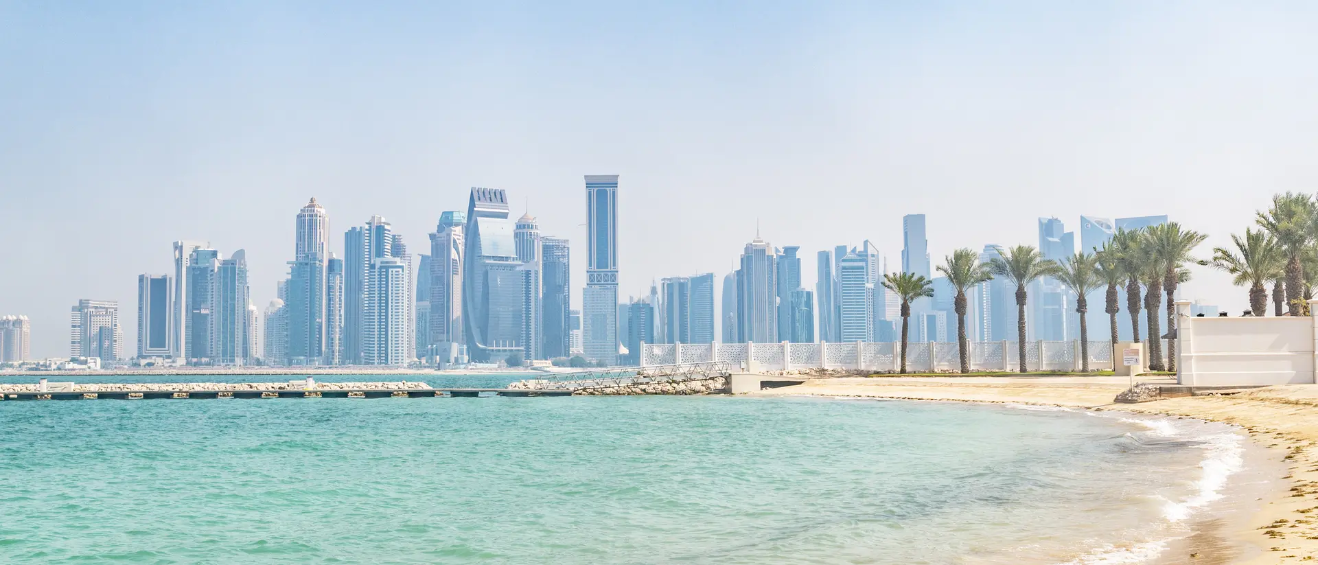 View of a sandy beach with palm trees and a skyline of tall buildings in the background under a clear sky.