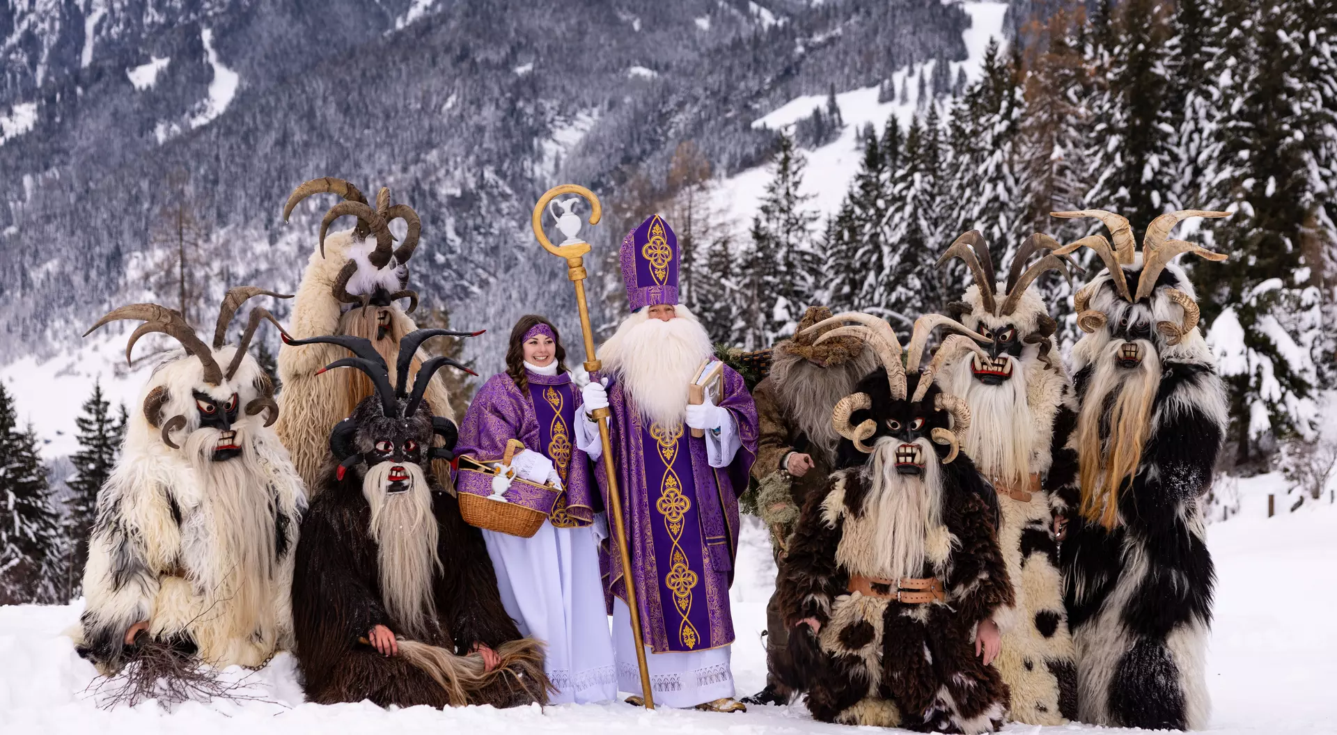 A snowy landscape featuring characters in costumes, including a robed figure and several horned, furry beings.