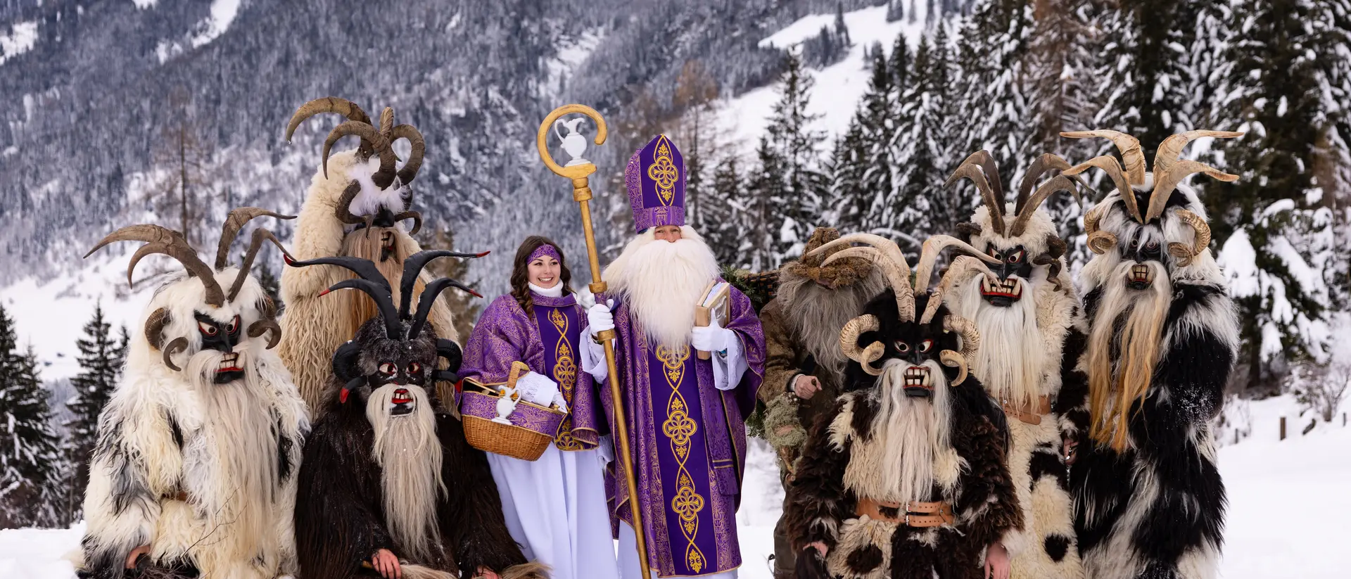 A snowy landscape featuring characters in costumes, including a robed figure and several horned, furry beings.