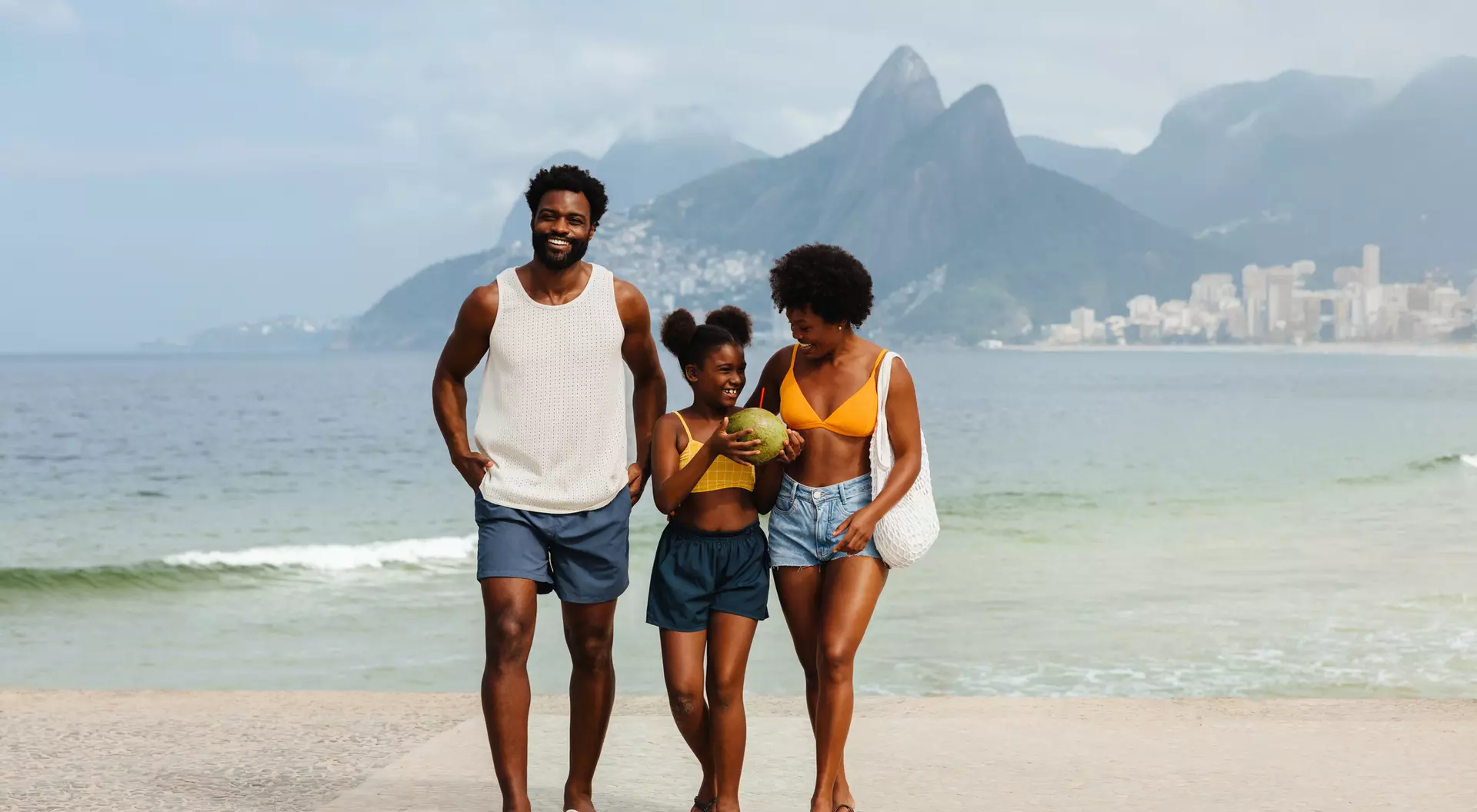 A smiling family walks by the beach, with mountains and a cityscape in the background.