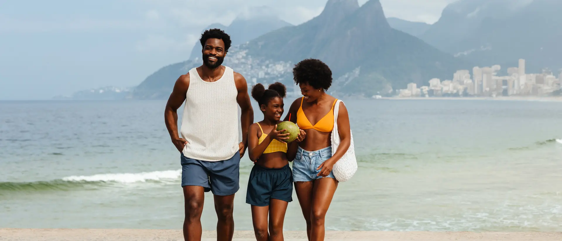 A smiling family walks by the beach, with mountains and a cityscape in the background.