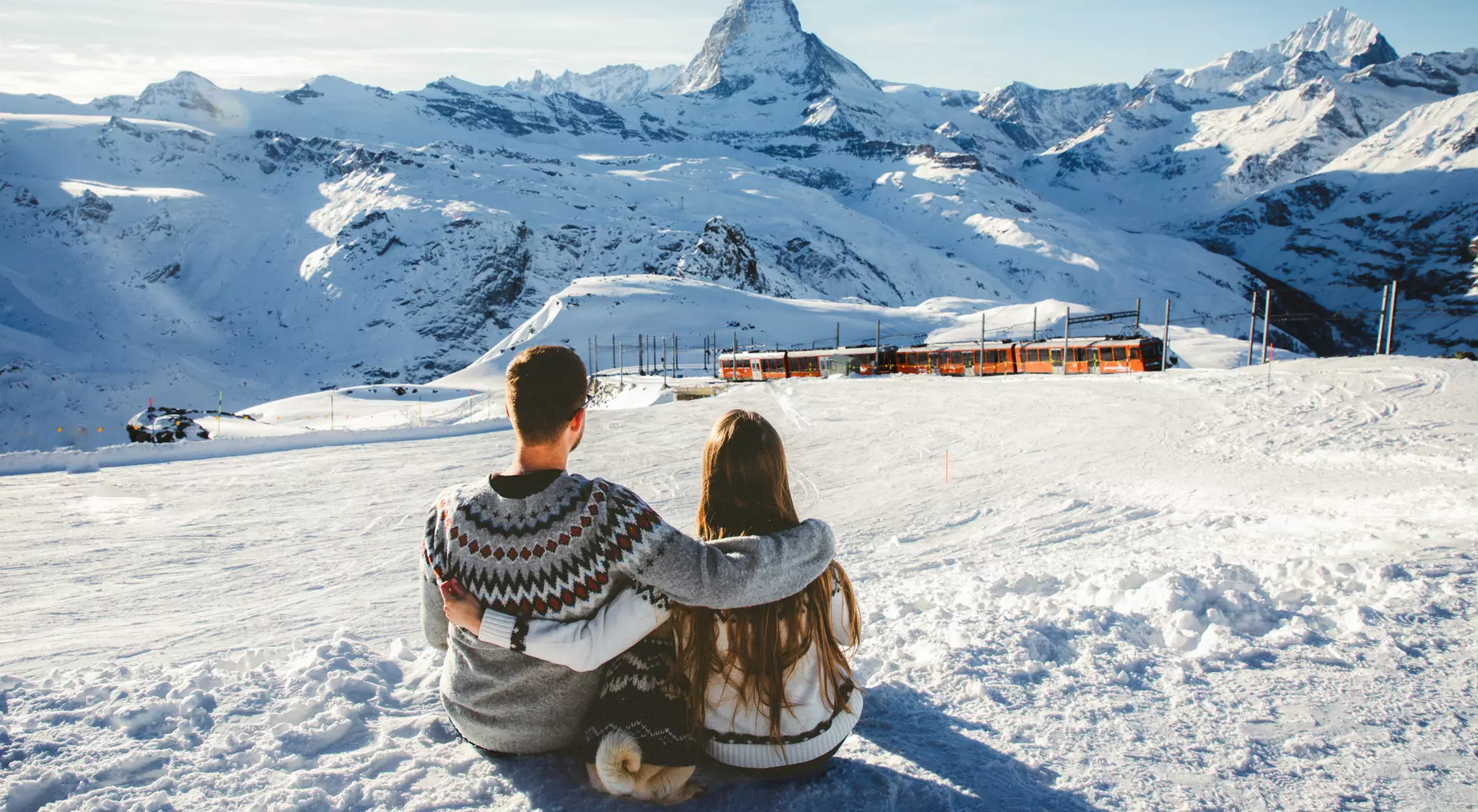 A couple sits together on a snowy slope, overlooking mountains, with the Matterhorn peak in the background.