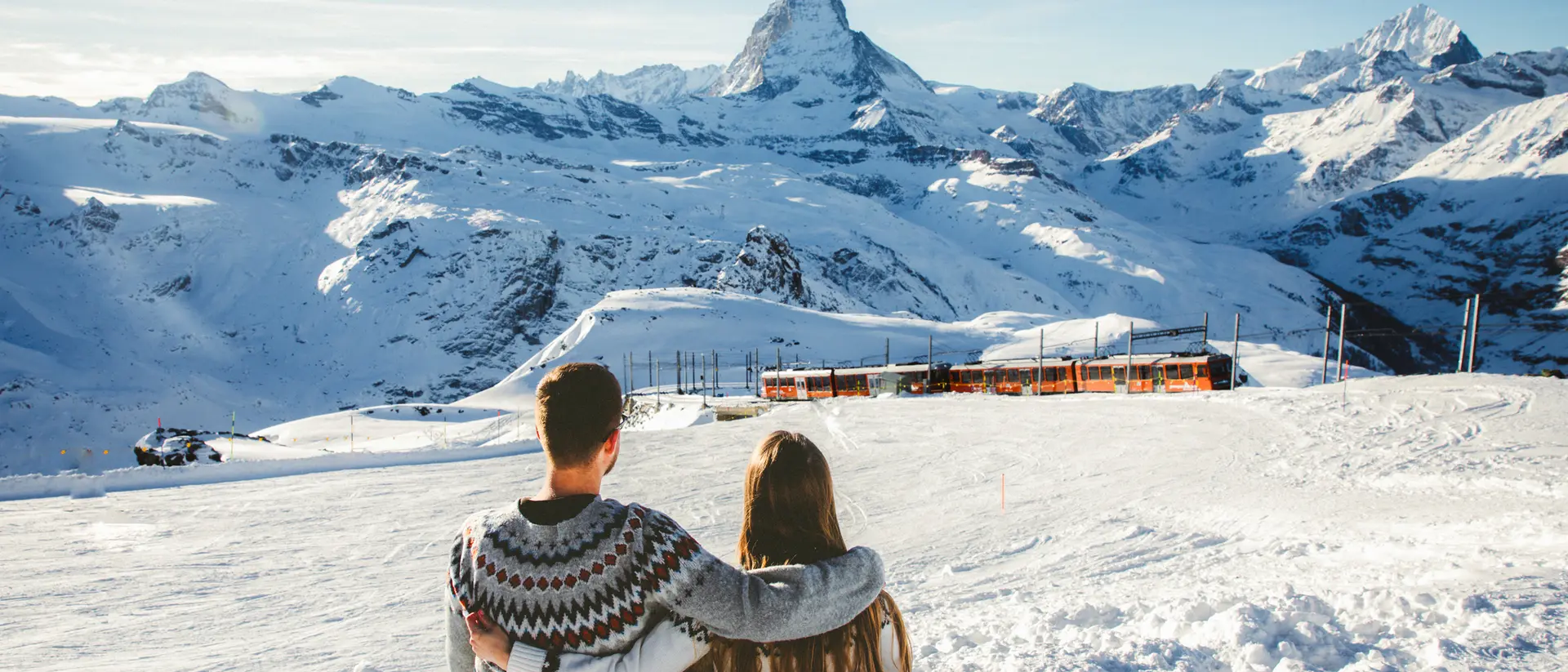 A couple sits together on a snowy slope, overlooking mountains, with the Matterhorn peak in the background.
