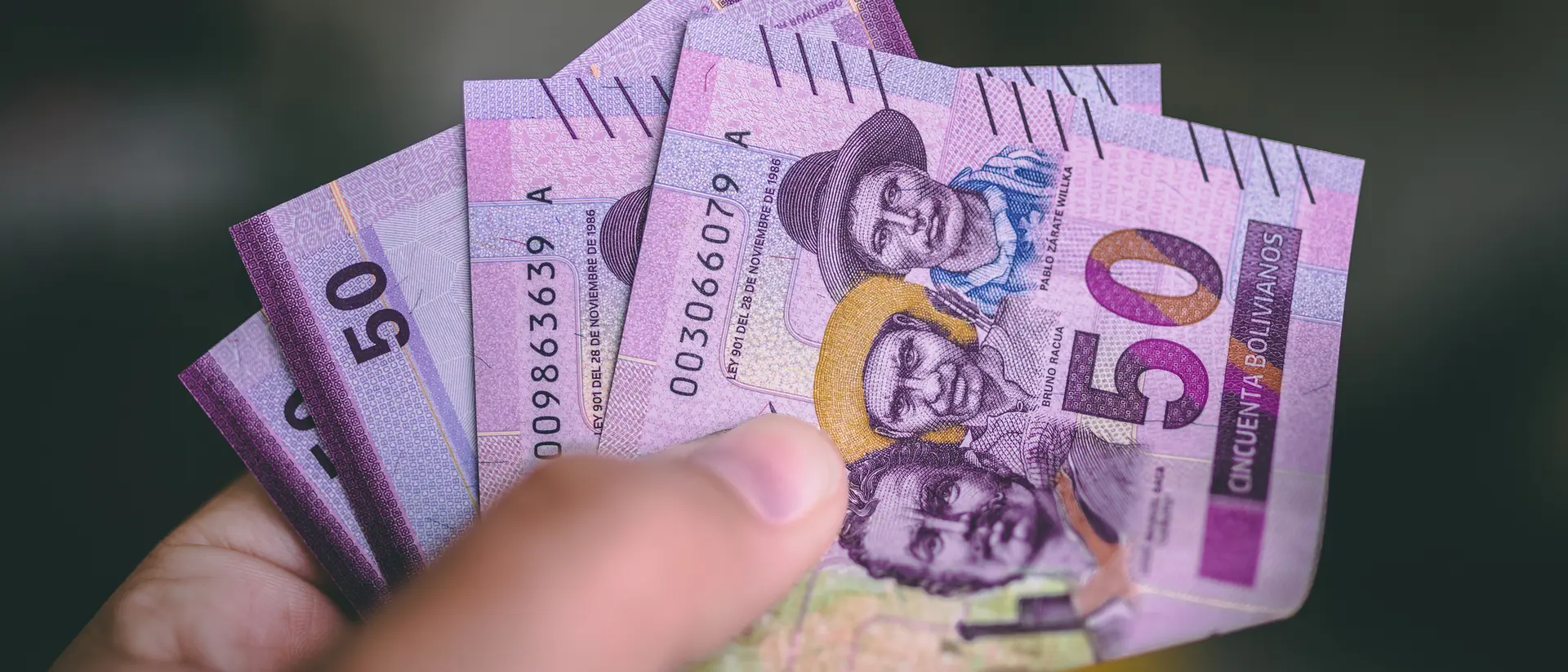 A hand holds several 50-unit banknotes featuring portraits of people, with purple and green designs.