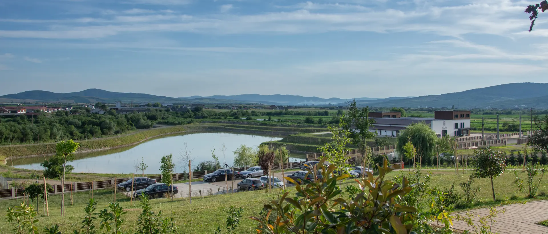 A scenic view of a pond, green fields, and mountains under a blue sky, with a modern building and parked cars in the foreground.