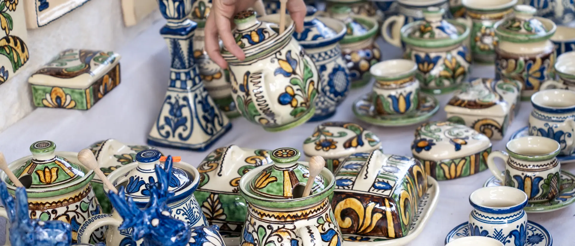 A hand reaches for a colorful, ornate pottery jar among various other decorated ceramics on a display table.